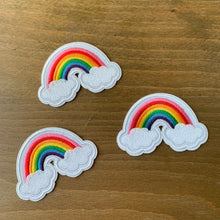 Load image into Gallery viewer, Three-pack 2 x1 inch embroidered iron-on patches with classic rainbows and two clouds, featuring a cute 80’s style. Ideal for customizing jackets, bags, shoes, and children's clothing. Shown on wood table.