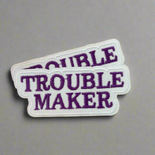 Load image into Gallery viewer, Close-up of two Trouble Maker Embroidered Iron On Patches in Purple and White Script, Displayed on a wood table.