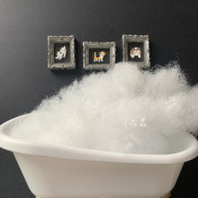 Load image into Gallery viewer, White faux fur or fleece stuffing piled in the basin of a small white bathtub, with three small framed dog portraits placed beside it on a dark background.