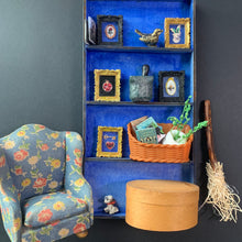 Load image into Gallery viewer, A miniature dollhouse-style room scene featuring a blue shelving unit decorated with small framed pictures, a bird figurine, and a basket, alongside a floral armchair and a round wooden box.