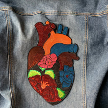 Load image into Gallery viewer, A close-up of a large iron-on patch shaped like an anatomical heart, featuring vibrant multicolored sequins and embroidered floral details on a black fabric base. Displayed on denim jacket.