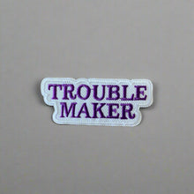 Load image into Gallery viewer, Close-up of Trouble Maker Embroidered Iron On Patch in Purple and White Script, Displayed on a wood table.