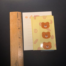 Load image into Gallery viewer, A close-up of a yellow patterned sticker strip of three brown bear faces and small pastry motifs, placed on top of a white lined sheet with a wooden ruler below it on a dark surface.