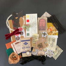Load image into Gallery viewer, A flat-lay collection of vintage-themed scrapbooking paper, stickers, ribbons, and craft embellishments arranged on a dark gray sheet of paper.