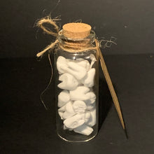 Load image into Gallery viewer, A small clear glass vial with a cork stopper, filled with many small white carved prop teeth, tied with twine, and photographed on a dark background. A kraft paper tag rests against the side of the bottle.
