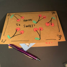 Load image into Gallery viewer, A handmade brown kraft paper card with a hand-drawn design of red cherries with green leaves and the text 'YOU'RE SO SWEET!', shown on a dark surface next to a purple pen.
