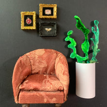 Load image into Gallery viewer, A miniature dollhouse-style armchair in rust pink damask fabric, positioned next to a white cylinder vase holding bright green coral-like decor, against a dark gray wall with three small framed art pieces.
