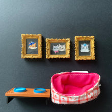 Load image into Gallery viewer, A dollhouse miniature accessory set featuring three small gold-framed picture frames with cat illustrations, a pink and white gingham check hat, and a brown stand with blue circular details, all arranged on a dark gray background.
