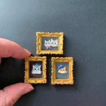 Load image into Gallery viewer, Three miniature gold-framed enamel pins featuring cartoon cat illustrations, held by two hands against a dark gray background.
