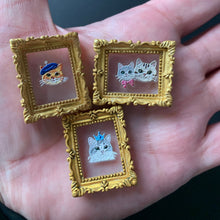 Load image into Gallery viewer, Three small enamel pin charms with ornate gold frames are held in a person's hand. Each pin features a white cat face on a pink background with different colored accents.
