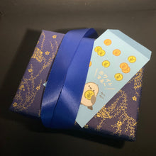 Load image into Gallery viewer, A small wrapped gift box with dark purple and gold floral-patterned paper, tied with a royal blue satin ribbon, with a light blue card featuring cartoon characters and gold coin illustrations peeking out. The box is photographed inside a dark box against a dark background.
