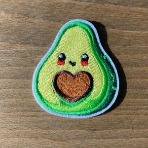 A photo of a small embroidered iron-on patches, about 1.85 inches tall: a bright green avocado with a heart-shaped pit featuring cute facial expressions.