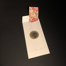 Load image into Gallery viewer, A white envelope or card holder on a black background, holding a round metallic coin and a small red patterned card or tag at the top.
