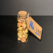 Load image into Gallery viewer, A small clear glass bottle with a cork stopper filled with tea-stained prop teeth, tied with twine and attached to a cardboard tag marked 'No. 133' in blue marker, set against a dark gray background.

