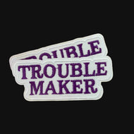 Close-up of two Trouble Maker Embroidered Iron On Patches in Purple and White Script, Displayed on a wood table.