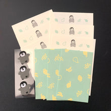 Load image into Gallery viewer, A set of cute penguin-themed stationery items, including cream-colored cards with gray and black penguin illustrations, mint green cards with yellow fruit prints, and a sheet of matching penguin stickers, all arranged on a dark background.
