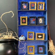 Load image into Gallery viewer, A blue wooden shelf display holding eight small framed art pieces with gold and black frames, featuring floral and symbolic illustrations. The shelf is styled with a black cauldron, string, a small blue pumpkin, and a broom against a dark gray background.
