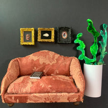 Load image into Gallery viewer, A miniature dollhouse scale sofa in a dusty rose and orange floral pattern, shown with a small book on its seat, set against a dark gray wall with three framed pictures and a green plant in a white pot.
