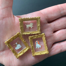 Load image into Gallery viewer, Three small square framed earrings displayed on an open hand. Each earring has a gold ornate frame, a pale pink translucent center, and a small white dog figure with a tiny orange accent.

