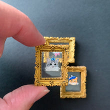 Load image into Gallery viewer, A close-up of two miniature gold picture frames being held by fingers against a dark gray background. The frames contain tiny illustrated scenes, including a white cat and a small dog.
