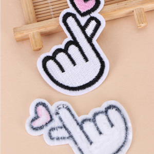 Two white embroidered Korean finger heart patches with pink hearts arranged on a bamboo tray. one is shown in reverse to highlight iron on.