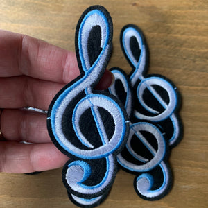 Large black and white embroidered treble clef iron-on patch in white, blue, and black, measuring 3.7 inches x 1.6 inches. Displayed on a wood surface.