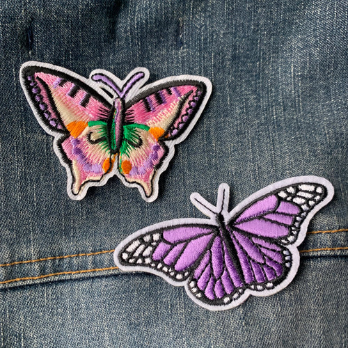 Two embroidered butterfly patches sewn on denim fabric, one large with a multicolored design and one small with a purple and black design.