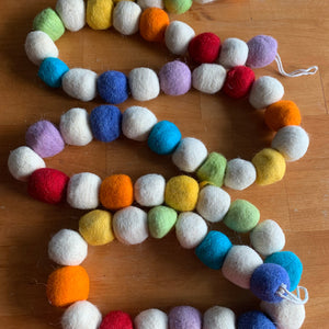 Colorful felt ball garland with soft, round felted wool balls in shades of red, orange, yellow, green, blue, purple, and cream, coiled neatly on a wooden surface.