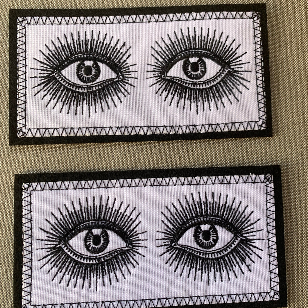 Embroidered Black and White Rectangle Vintage-look eyes iron-on patch, close-up on beige fabric background.