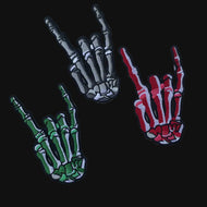 Set of three embroidered iron-on skeleton hand patches—gray, red, and green—each making the rock-on / I-love-you sign on a neutral background.